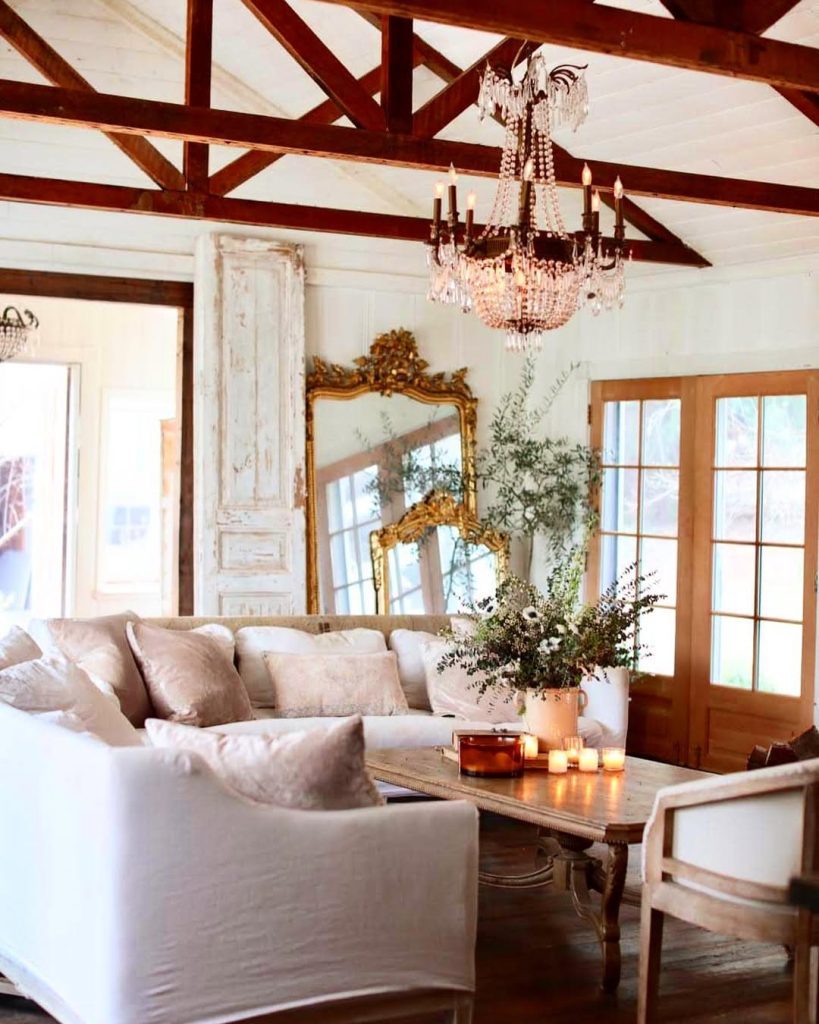 French country living room with exposed beams, crystal chandelier, neutral sofas, and ornate mirrors