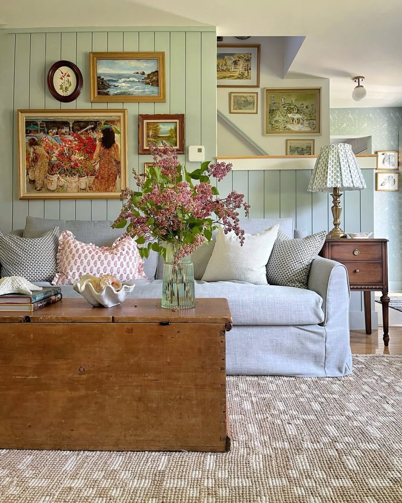 Cottage living room with sage green paneling, vintage paintings, blue furniture, and fresh flower arrangements