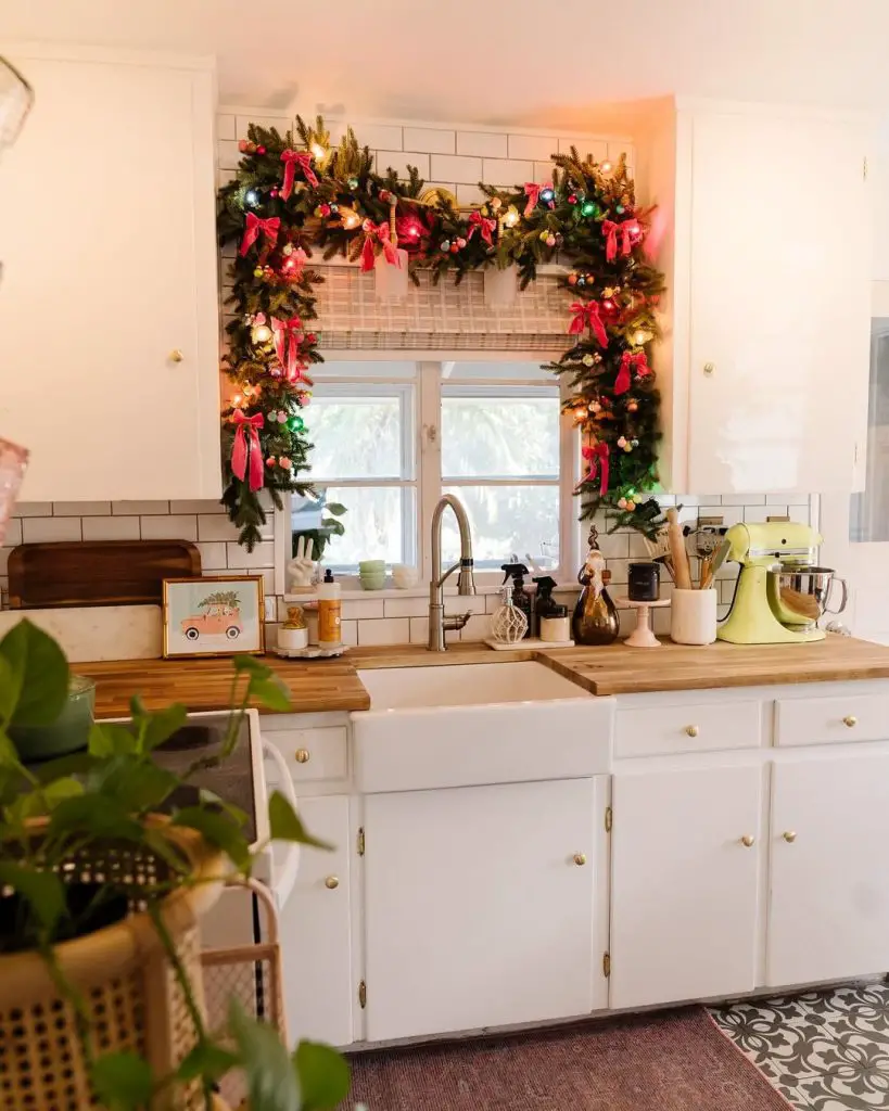 Kitchen window decorated with evergreen garland, red ribbons, twinkling lights, white cabinets